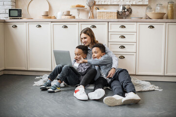 Happy multinational family of mother and two sons sitting on warm floor in light kitchen during...