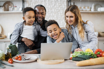 Multinational family of african cute little sons, father and caucasian mom ordering ingredients for holiday meal using laptop, sitting at wooden table in kitchen, having fun, enjoying weekend.