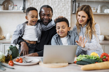 Multinational family of african cute little sons, father and caucasian mom ordering ingredients for holiday meal using laptop, sitting at wooden table in kitchen, having fun, enjoying weekend.
