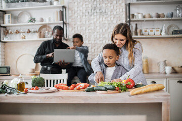 Pretty mother and son preparing tasty food at kitchen. Mommy teaching lovely kid to cook while happy father and child boy spending time using laptop computer. Healthy meal and dinner preparation.