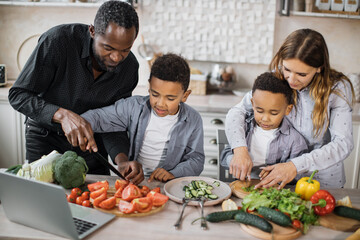Happy young parents teaching little preschooler boys to chop vegetables with knife preparing salad for lunch together loving mom, dad and small sons kids cooking dinner together.