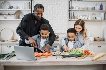 Smiling african cute little sons and their beautiful young parents having online cooking class on laptop together slicing fresh vegetables with knife and making salad, food in kitchen at home.