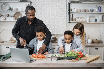 Smiling african cute little sons and their beautiful young parents having online cooking class on laptop together slicing fresh vegetables with knife and making salad, food in kitchen at home.