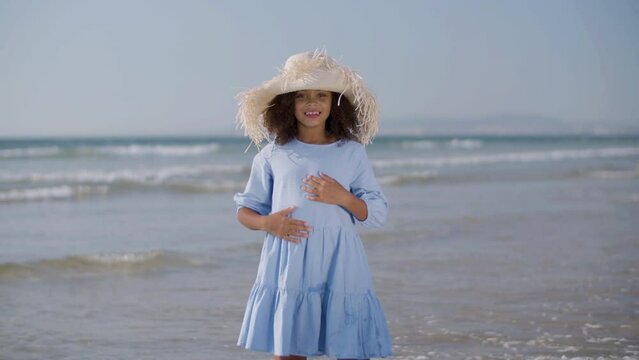 Happy Black girl in straw hat dancing on the beach as waves rushing to the shore in background. Smiling little girl in blue dress enjoying holiday time at seaside. Childhood, vacation concept.