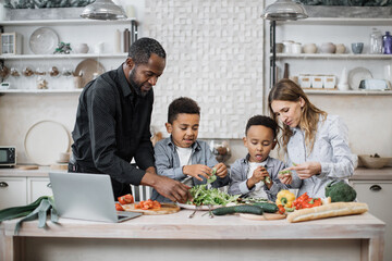 Cute boys helping their young parents tearing freshly washed, fresh clean leaves of lettuce into bowl, preparing healthy salad. Multinational mom and dad enjoying cooking healthy food with little sons