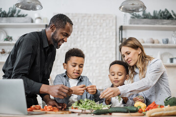 Multinational family cooking healthy salad on light kitchen in weekend. Cute boys helping their young parents tearing freshly washed, fresh clean leaves of lettuce into bowl, preparing healthy salad.