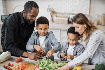Close up view of parents and their sons cooking in kitchen preparing food salad together. Happy boys children assist mom and dad, helping with dinner or lunch, tearing leaves of lettuce into bowl.