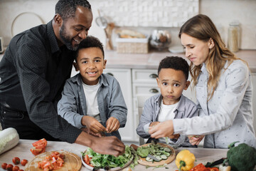 Naklejka premium Closeup of smiling boys children helping mom and dad with dinner or lunch, tearing leaves of lettuce into bowl. Multinational parents and their sons cooking in kitchen preparing food salad together.