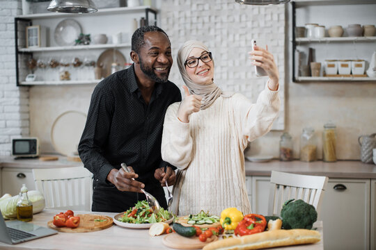 Cheerful African Man And Muslim Woman Smiling And Showing Thumb Up While Making Video Call On Smartphone And Cooking Delicious Salad From Fresh Vegetables In Modern Kitchen.