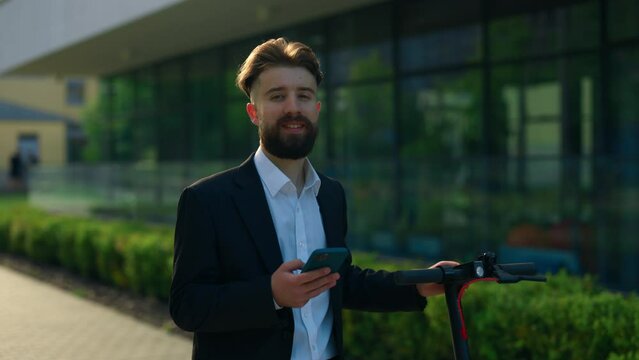 Portrait of the business man with smartphone in his hand, wearing in formal suit, carrying the electric bicycle. Male manager looking at camera standing near office centre.