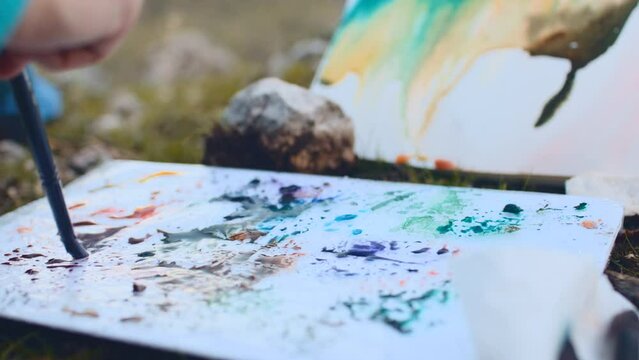 Close-up of a girl sitting outdoors and mixing colors on a color palette with brushes, for art, education, creativity and education concept.
