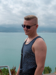 A handsome adult man with a stylish hairstyle in sunglasses on the seashore in a T-shirt smiles looking into the frame