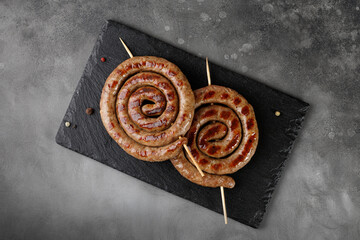Grilled pork sausages with sauce on black stone board on dark background. BBQ. Homemade Snail round sausage. Top view, copy space. - obrazy, fototapety, plakaty