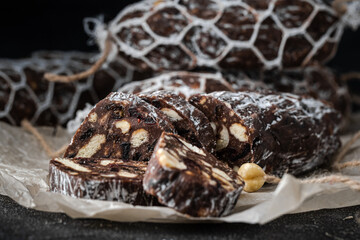 Chocolate salami filled with almonds, hazelnuts and raisins on the black background. Italian sweet sausage shaped dessert sprinkled with powdered sugar. Christmas and New Year festive pastry