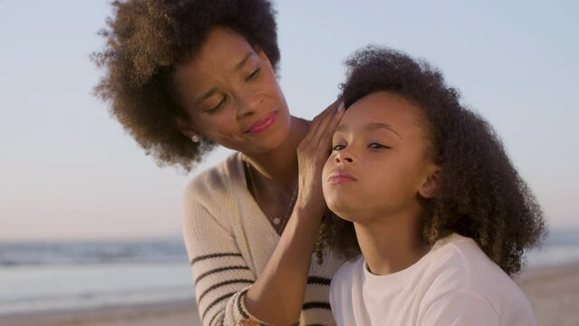 Medium closeup of mum and daughter having picnic at beach. Caring Black woman brushing off crumbs on childs face while cute girl chewing, eating up snack. Slow motion. Parenthood, motherhood concept.