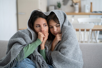 Unhappy frozen woman with daughter are sits on couch cuddling up to each other wrapped in warm...