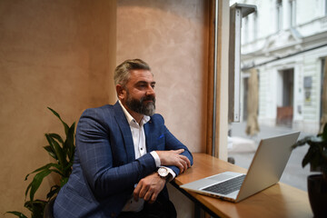 Modern senior businessman sitting in cafe and using laptop and smartphone