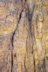A male rock climber in a red t-shirt climbs a route on a steep granite wall on the rocks of the mountains