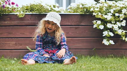 A little girl in a panama hat sits by the white petunias in the garden. - obrazy, fototapety, plakaty