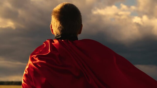 Little hero in red cloak looks at approaching storm. Bold child winner in red raincoat, nature, Boy Plays Superhero in red cape, Childrens dream. Happy child playing super hero in front of blue cloud