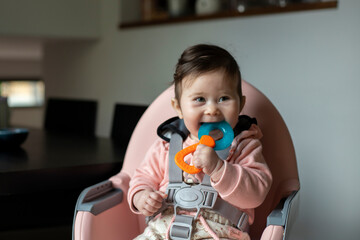 Nine-month-old cute baby sitting in pink baby chair, smiling and biting blue-orange teether in hand.