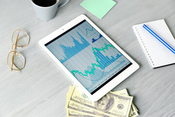 Modern tablet computer with stock data, dollar banknotes, eyeglasses and notebook on grey wooden background. Finance trading