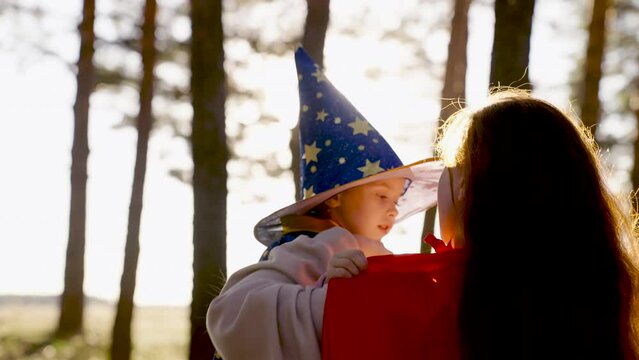 Child plays in mantle of magician, mother in red cloak. Childhood dreams, fantasies to become wizard, Child plays with his mother in costumes of wizard, playing in park on Halloween. Happy family.