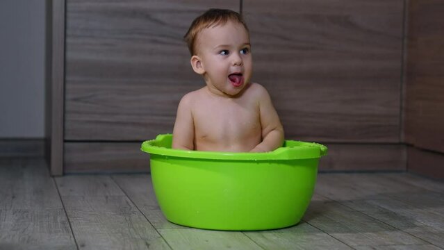 Sweet funny kid sitting naked in the washbowl making faces and showing tongue. Adorable baby having fun during wash time.