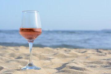 Glass of tasty rose wine on sand near sea, space for text