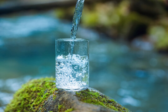 Fresh water pouring into glass on stone with moss near river. Space for text © New Africa