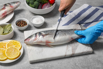 Woman cutting fresh raw sea bass fish at light gray table, closeup