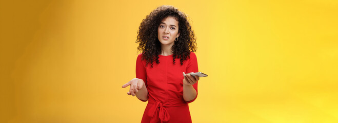 So waht, I confused. Questioned uncertain woman with curly hair in red dress shrugging looking clueless as holding hand cannot understand where order as checking mail box via device over yellow wall - obrazy, fototapety, plakaty