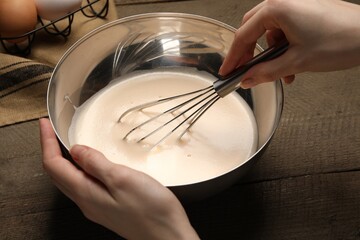 Fototapeta na wymiar Woman whipping ingredients with balloon whisk at wooden table, closeup. Cooking delicious eggnog