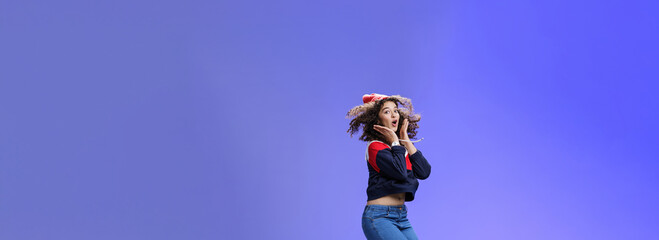Fototapeta na wymiar Surprised and amazed amused good-looking woman with curly hair in winter beanie and sweatshirt open mouth from joy and amazement jumping over blue background having fun feeling playful