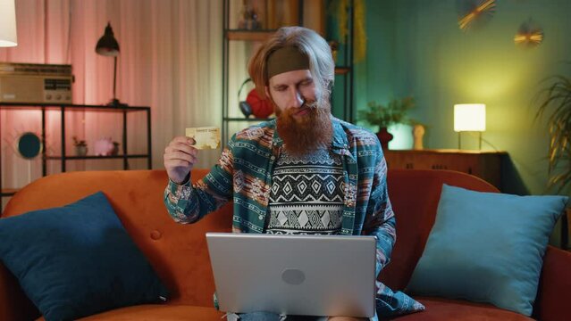 Bearded redhead man using credit bank card and laptop computer while transferring money, purchases online shopping, order food delivery at evening home. Young hippie guy on couch at home evening room