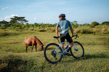 A young bearded cyclist is biking through a field with a horse