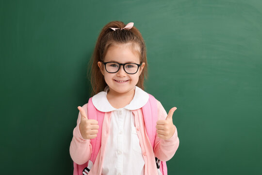 Happy little school child showing thumbs up near chalkboard