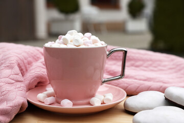 Cup of tasty cocoa with marshmallows, pink sweater and cookies on wooden table outdoors