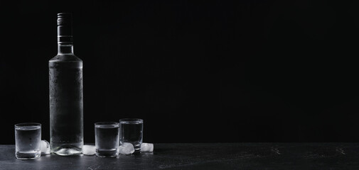 Bottle of vodka and shot glasses with ice on table against black background, space for text. Banner...