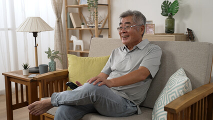 side view elderly Korean man hands console tv channel media sit on comfort sofa in house living room indoors. laughing old male enjoy funny talk show on television