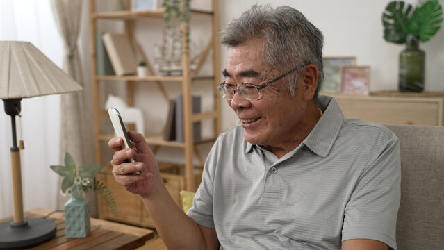 Laughing Mature 80s Man Sit On Sofa In Living Room Using Modern Smartphone Gadget. Joyful Senior Grandfather Rest On Couch At Home Browse Internet On Cellphone. Older People Technology Life Concept