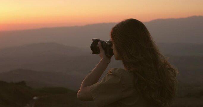 Back view of caucasian woman hand holding professional camera and taking photo on the red sunset at the camping in the mountains. Evening sky scene with golden light from the setting sun in summer