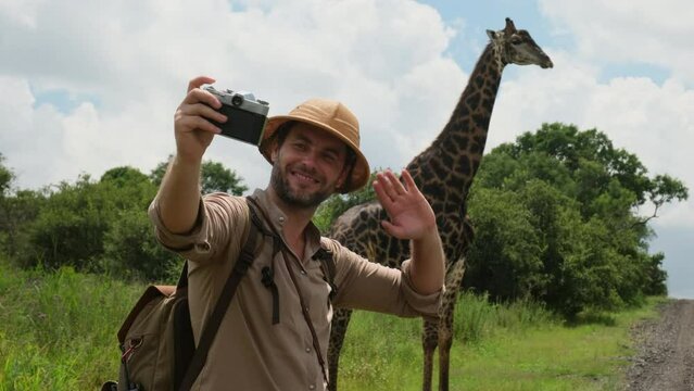 man Photographer on safari photographs a wild giraffe who eats leaves on a bush in the African savannah, in the dry season. male traveler in safari hat taking photo of giraffe taking a selfie