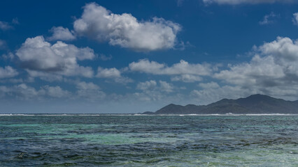 The boundless turquoise ocean glitters in the sun. Ripples on the water. In the distance, against the background of blue sky and clouds, the outlines of tropical islands. Seychelles