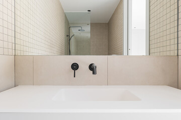 Modern bathroom with beige tiles, white sink and black faucet. Shower zone with glass partition reflected in the mirror