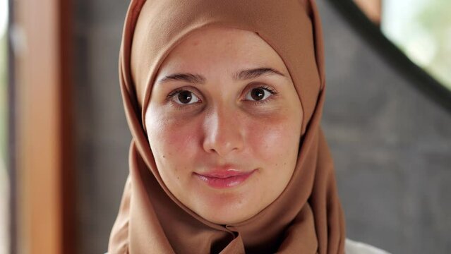 Positive woman with hair covered by hijab look straight to camera and smile, then smile slightly more and we see dental braces on her teeth. Headshot of young lady wearing traditional headcovering