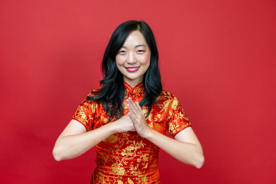 Asian Chinese Woman With Red Cheongsam Or Qipao Doing Polite Respectful Gesture For Wishing The Good Luck And Prosperity In Chinese New Year Celebration Holiday Isolated On Red Background Concept