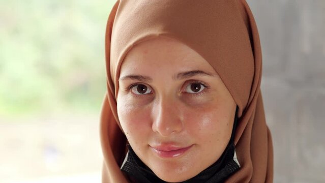 Young woman with head and hair wrapped in hijab wearing black surgical mask. She look aside, then pull mask down and look straight to camera. Portrait shot with blurred background