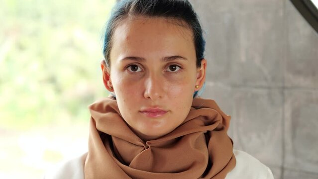 Young woman with a serious face looks straight into the camera, pull down her hijab and we see her hair dyed blue. Portrait shot of strong lady.