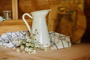 Rustic kitchen items. Enameled metal white jug for milk or water and a bouquet of wildflowers on the kitchen table in a log house
